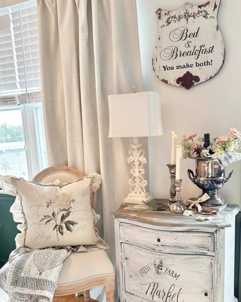 A cozy corner with a chair, floral pillow, lace throw, distressed white nightstand, lamp, silver vase with flowers, candles, and a decorative “Bed & Breakfast” wall sign, perfect for those who love French country decor.
