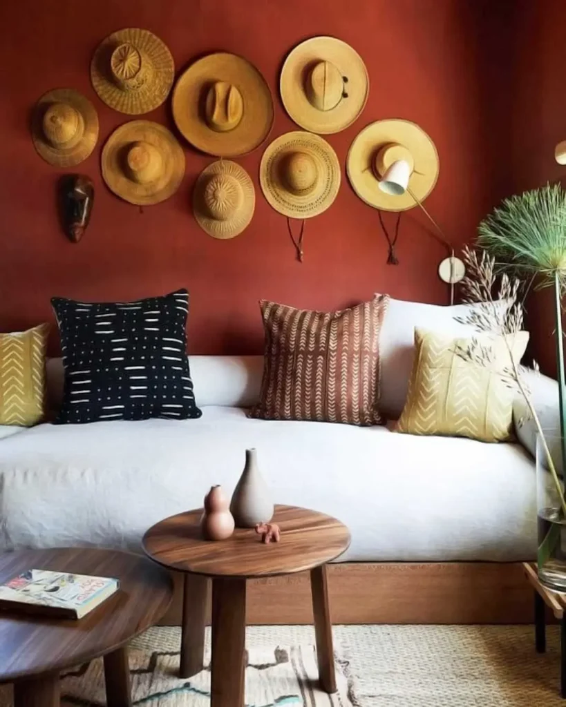 A white sofa with patterned pillows sits against a rust-colored wall decorated with a woven hat collection. Two wooden side tables hold small vases and a magazine, while tall dried plants stand in the corner.