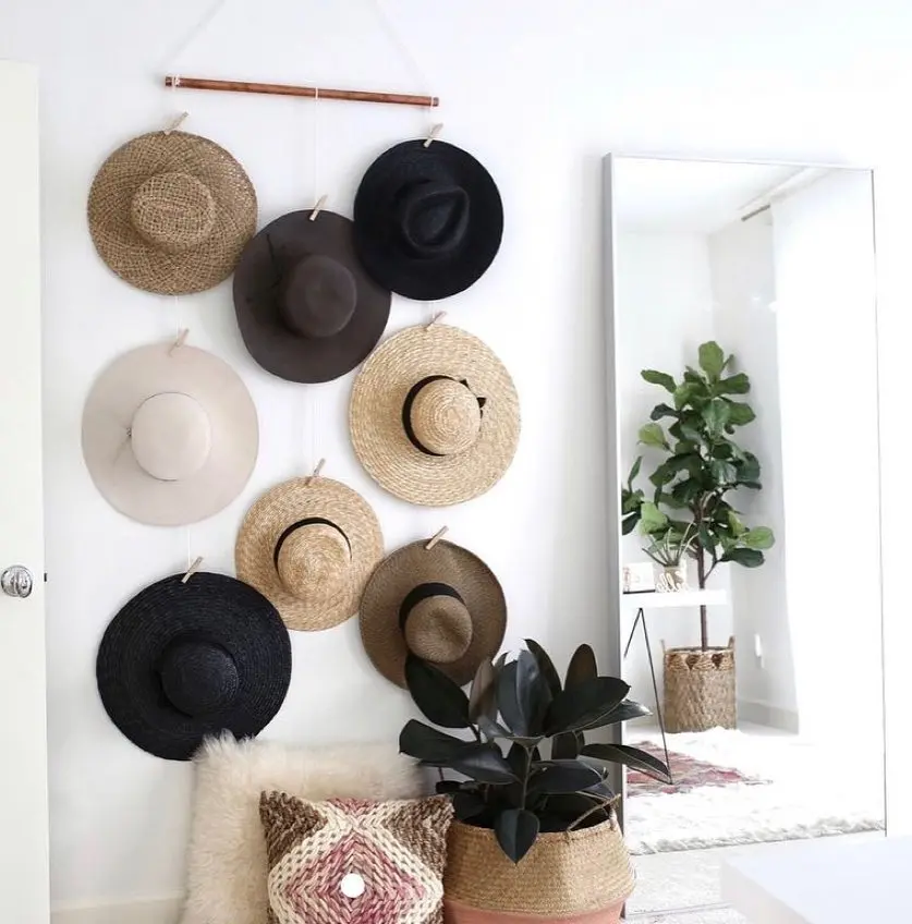 Eight stylish hats are displayed on a white wall above a plant and a patterned cushion, with a large mirror and a potted tree visible in the background. This elegant hat wall arrangement adds character to the space.