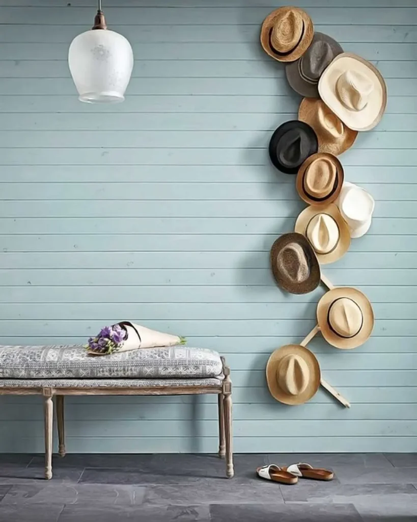 A light blue wooden wall features a hat gallery wall with various hats arranged vertically, a small bench holding a bouquet, and a pair of shoes on the grey tiled floor.