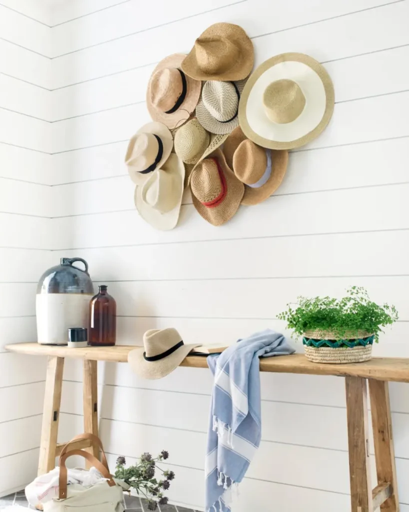 A wooden table with a blue-striped towel, a hat, two bottles, and a basket with greenery sits below eye-catching hat wall decor displayed on white shiplap.