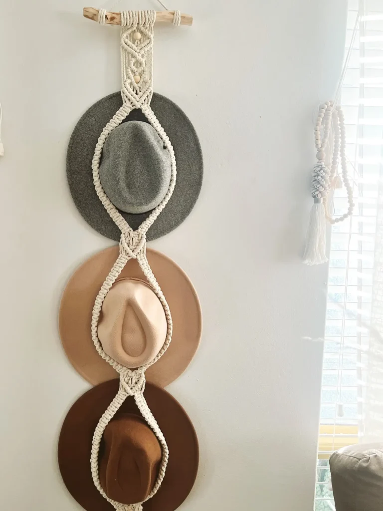 Three hats in gray, beige, and brown are displayed vertically on a wall, held by a macramé hanger next to a window with blinds.