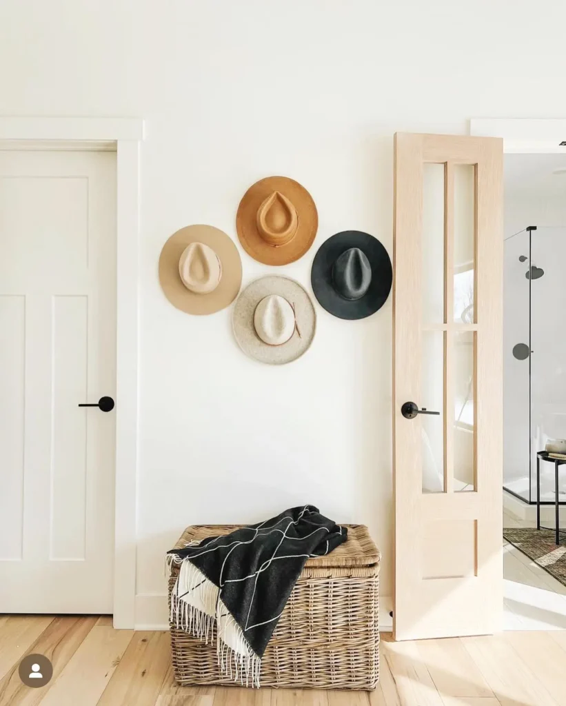 Four stylish hats in neutral tones hang on a white wall above a wicker basket with a folded black and white blanket, next to two doors in a bright, minimal entryway.