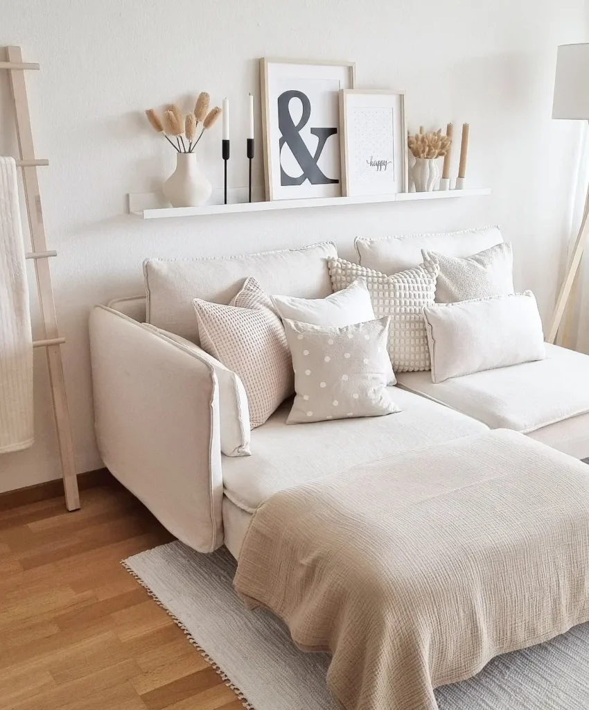 A hygge-inspired neutral-toned living room features a white sofa, assorted beige and white pillows, a light throw blanket, framed prints on a shelf, and minimalist decor atop a warm hardwood floor.