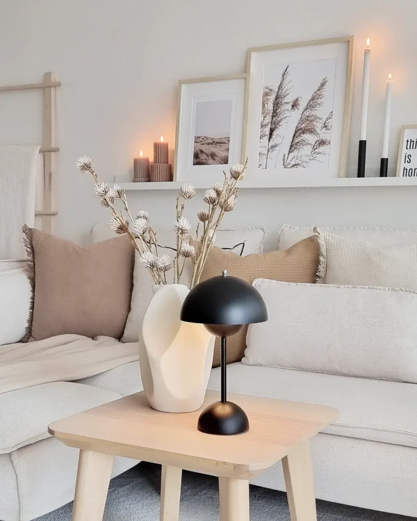 A modern living room with neutral-toned decor captures hygge winter vibes, featuring a table with a black lamp and vase, plus candles and framed art arranged on a shelf in the background.