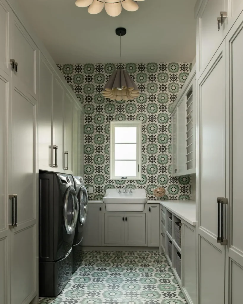 A narrow laundry room features patterned tile walls and floor, a washer and dryer, a farmhouse sink, cabinets, and overhead pendant lights.