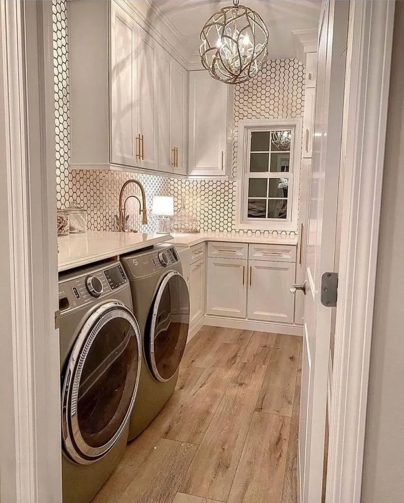 A modern farmhouse laundry room with a front-loading washer and dryer, white cabinets, gold fixtures, wood flooring, and hexagonal tiled walls, all illuminated by a stylish globe light fixture.
