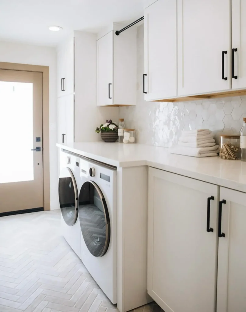 Modern farmhouse laundry room with front-loading washer and dryer, white cabinets, stacked towels, and a jar filled with clothespins on a crisp white countertop.