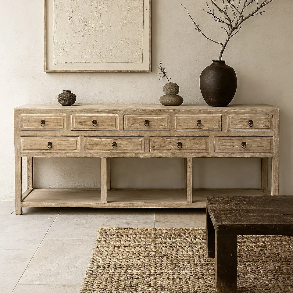 A rustic console with nine drawers stands against a neutral wall, topped with three vases and a branch; a textured rug and wooden table are in the foreground.