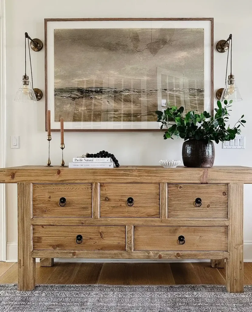 A rustic console with drawers stands against a wall, topped with books, candlesticks, a glass bowl, black beads, and a vase of greenery; artwork and wall sconces hang above.