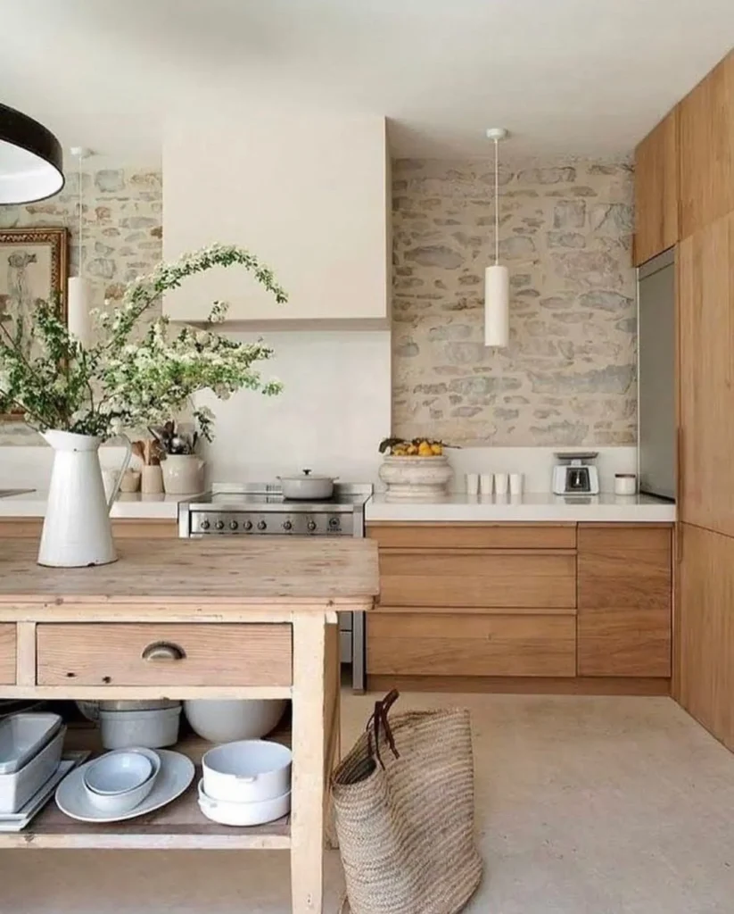 Modern kitchen with stone wall, wooden cabinets, a stove, and a wooden island. Rustic kitchen accessories, like a white pitcher with greenery and woven bag on the floor, add charm; open shelves hold dishes.