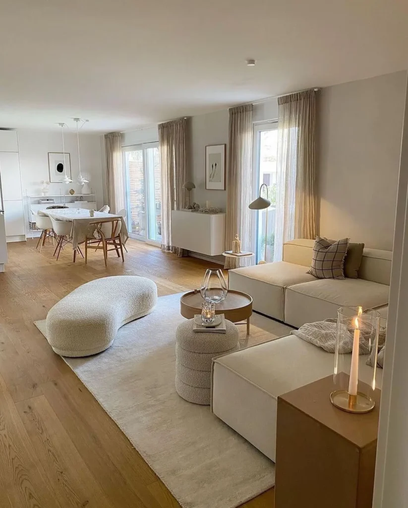 A modern, minimalist Scandinavian living and dining area with neutral tones, light wood flooring, a sectional sofa, abstract furniture, and large windows with sheer curtains.