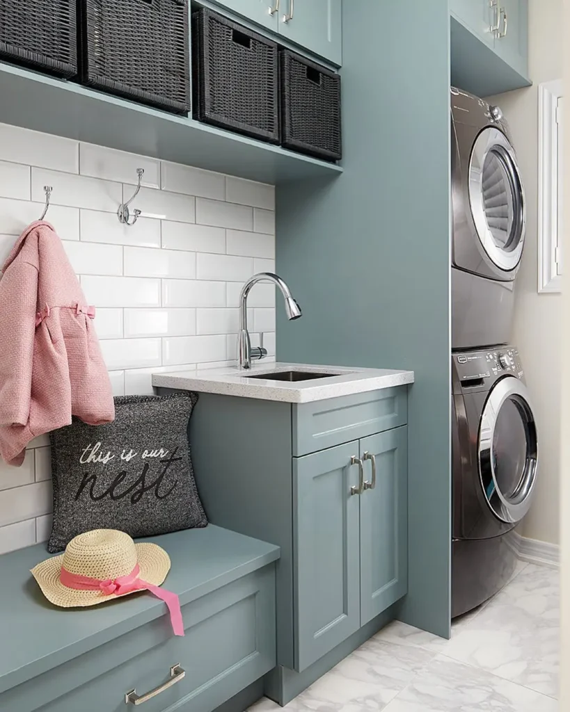 A small laundry mudroom featuring stacked washer and dryer, built-in sink, cabinets, black storage baskets, a bench with a hat and pillow, and a pink sweater hanging on a hook.