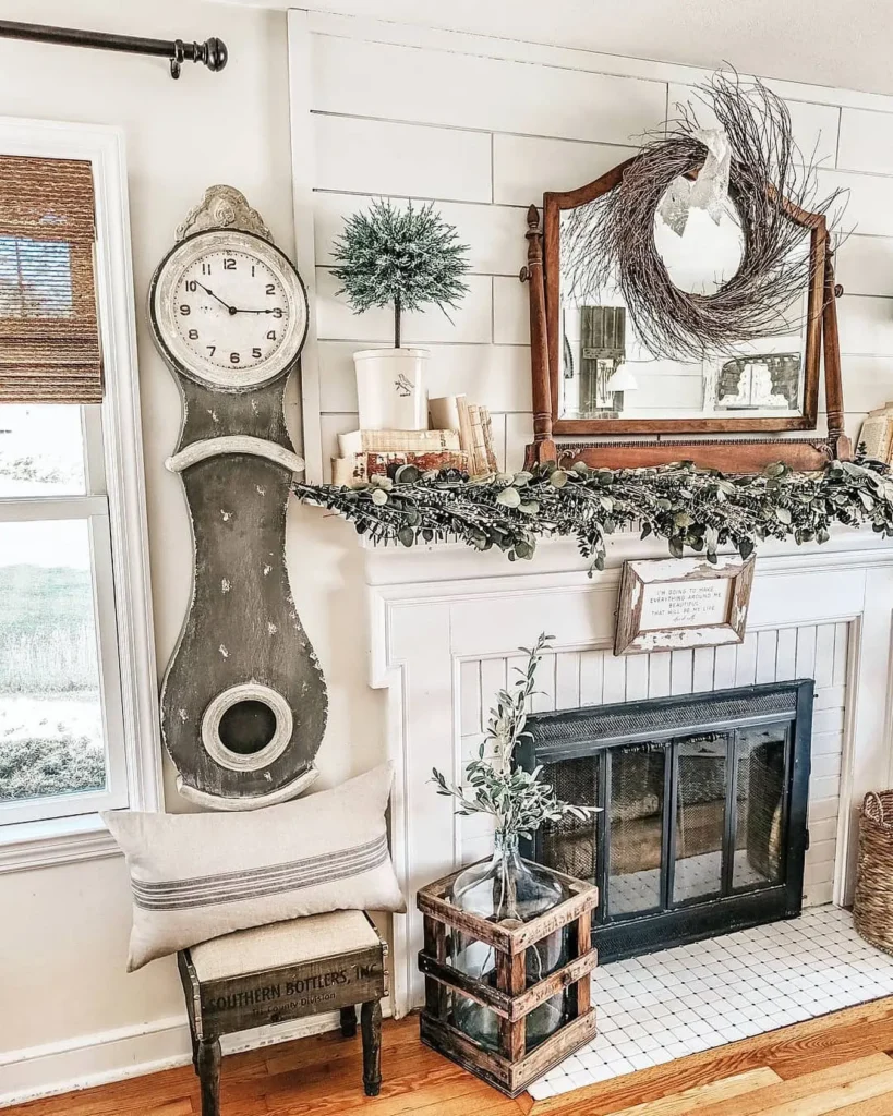 A rustic living room with a vintage clock, mirror with a wreath, potted plant, and a garland on the vintage farmhouse mantel above the fireplace adorned with decorative accents.