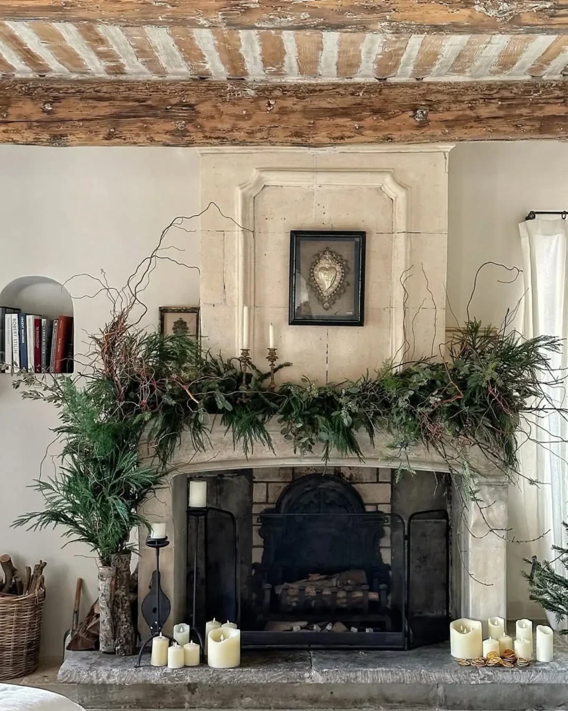A stone fireplace decorated with greenery and candles, featuring vintage farmhouse charm with a framed heart artwork above the mantle and rustic wooden ceiling beams overhead.