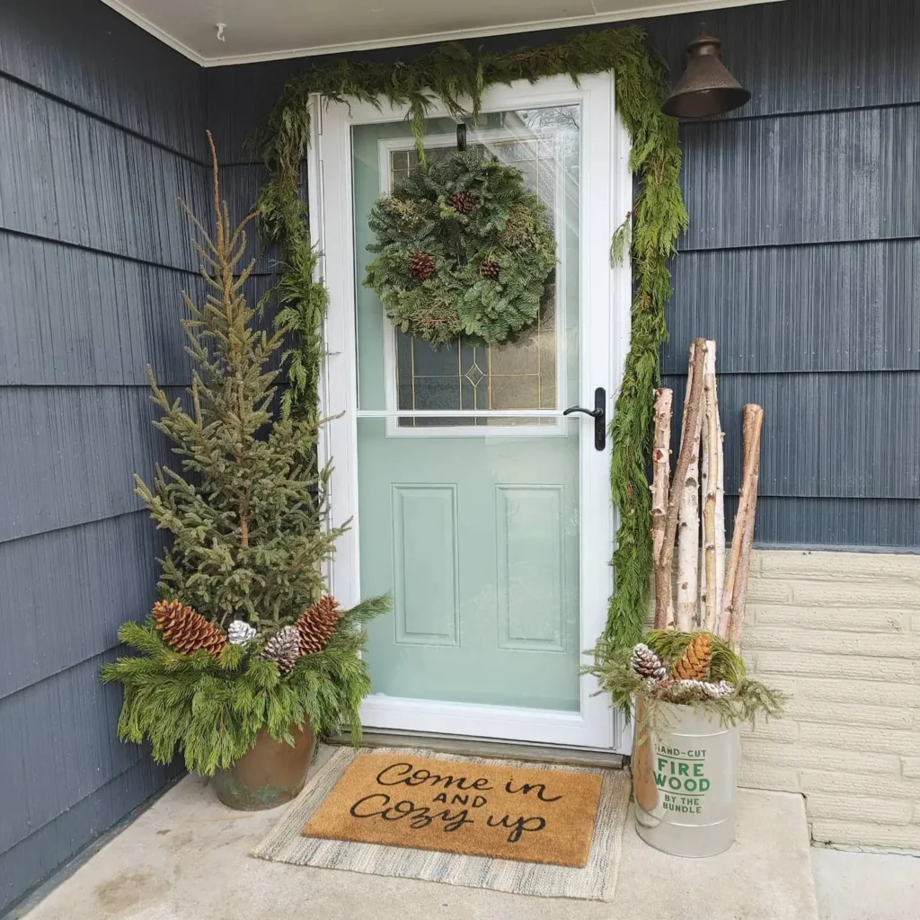 A winter front porch features a door adorned with a wreath, greenery, and birch logs; a mat reads “Come in and Cozy up.” To the left of the door, a potted plant filled with pinecones adds to the seasonal charm.