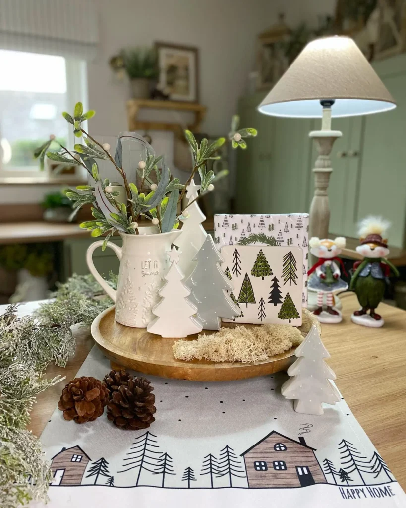 A wooden tray with ceramic trees, a white jug with greenery, pinecones, and holiday figurines decorates a cozy table set with a winter table runner in a warmly lit room.