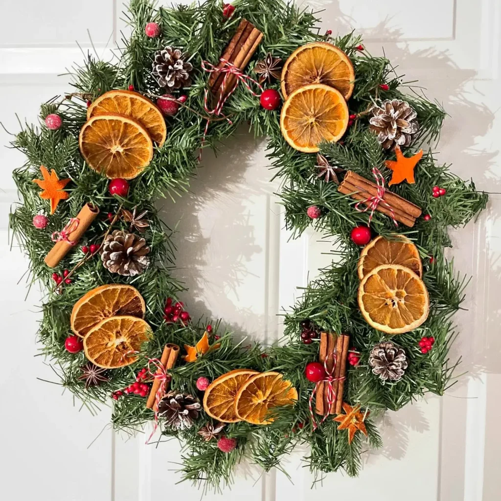 A festive winter wreath adorned with dried orange slices, pinecones, cinnamon sticks, small red berries, and orange star-shaped pieces hangs beautifully on a white door.
