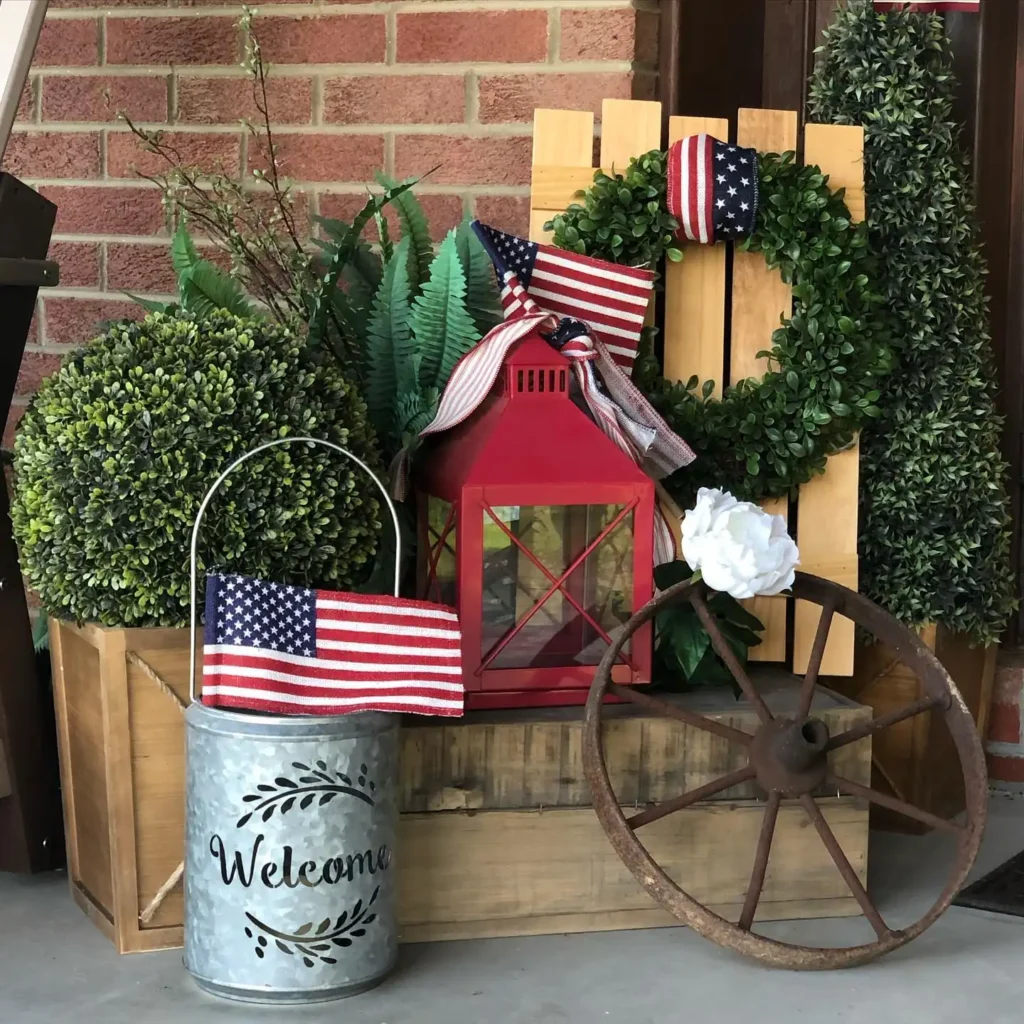 Fourth of July front porch decor vignette with American flags, greenery, a red lantern, rustic wood crate, and farmhouse-style patriotic accents.