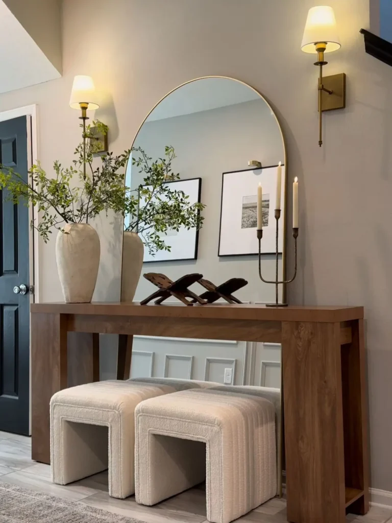 A modern wooden console table with a large round mirror, two white vases filled with branches, three candles, and two upholstered stools creates a chic look in this hallway.
