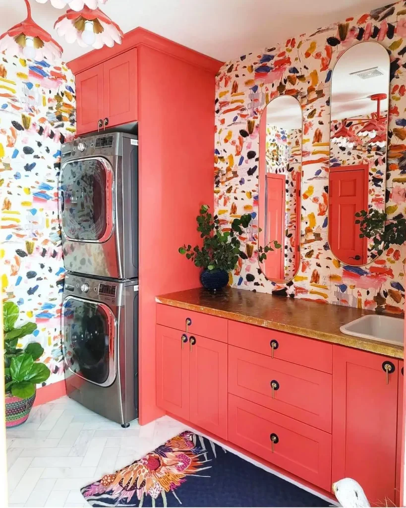 Bold laundry room with coral cabinets, abstract colorful wallpaper, stacked washer and dryer, arched mirrors, and gold countertop.