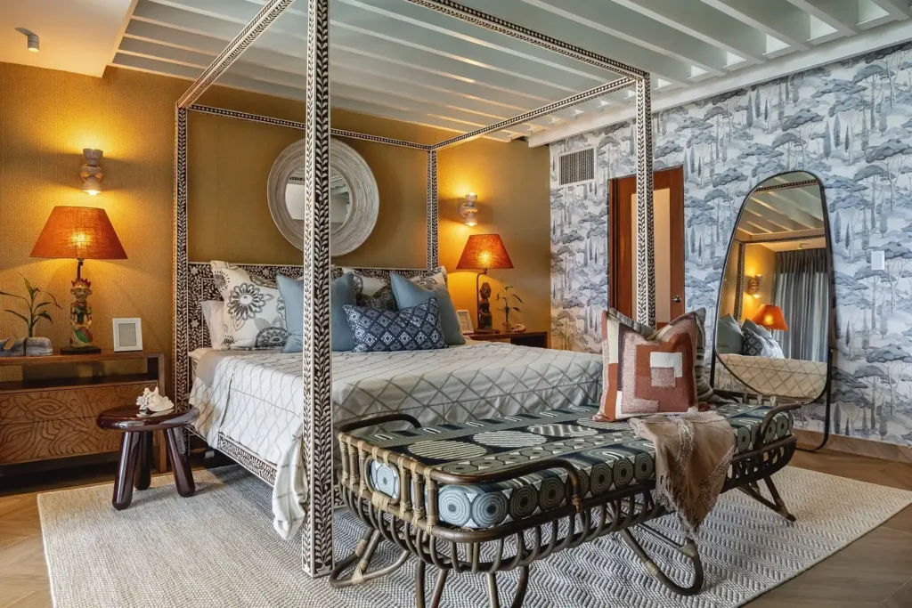Eclectic bedroom decor featuring a patterned four-poster bed, layered textiles, wallpaper, and warm ambient lighting.
