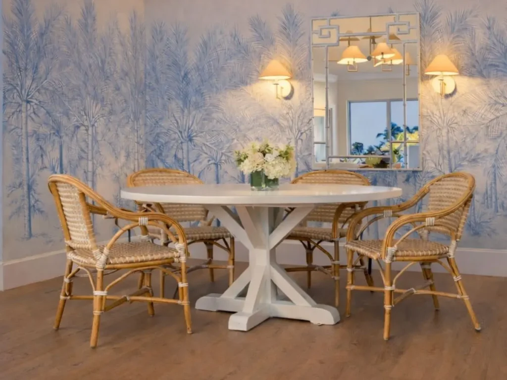 Coastal dining room styled with rattan dining chairs, a white pedestal table, palm tree wallpaper, and warm lighting, showcasing earthy and textured rattan decor.