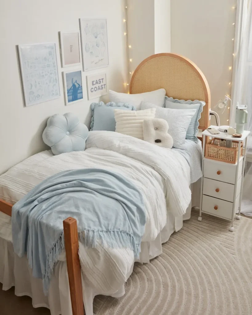 A neatly made single bed with white and light blue bedding, decorative pillows, framed wall art, string lights, and a small bedside cart create a cozy, well-lit dorm bedroom.