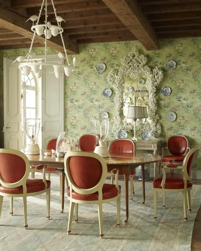 A dining room with a wooden table, six red and cream chairs, floral wallpaper in a Clay White & Willow Green color palette, ornate mirror, decorative plates, and a white chandelier under a wooden-beamed ceiling.
