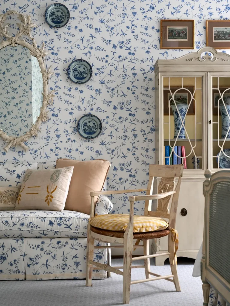Living room features a Cornflower Blue & Buttercream color palette with floral wallpaper, a white sofa, decorative plates, an ornate mirror, a yellow-cushioned wooden chair, and a vintage cabinet with china.