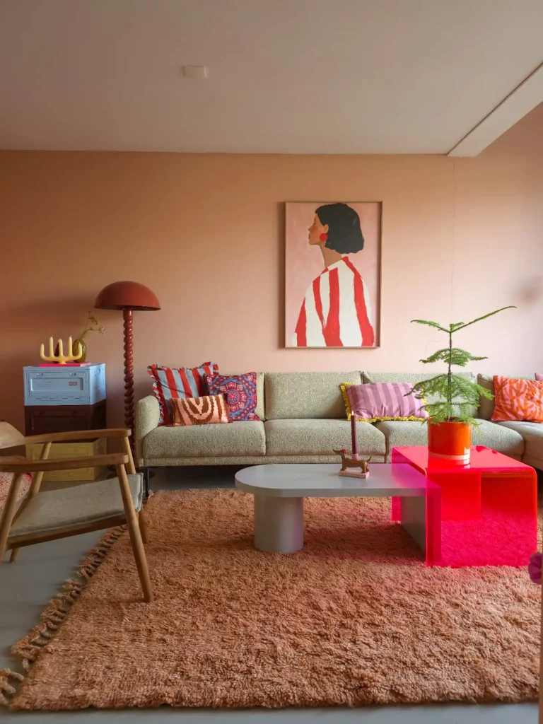 A modern living room with pastel walls, a beige sofa with funky colorful cushions, a red lamp, framed artwork, a coffee table, and a small potted plant on a brown rug.