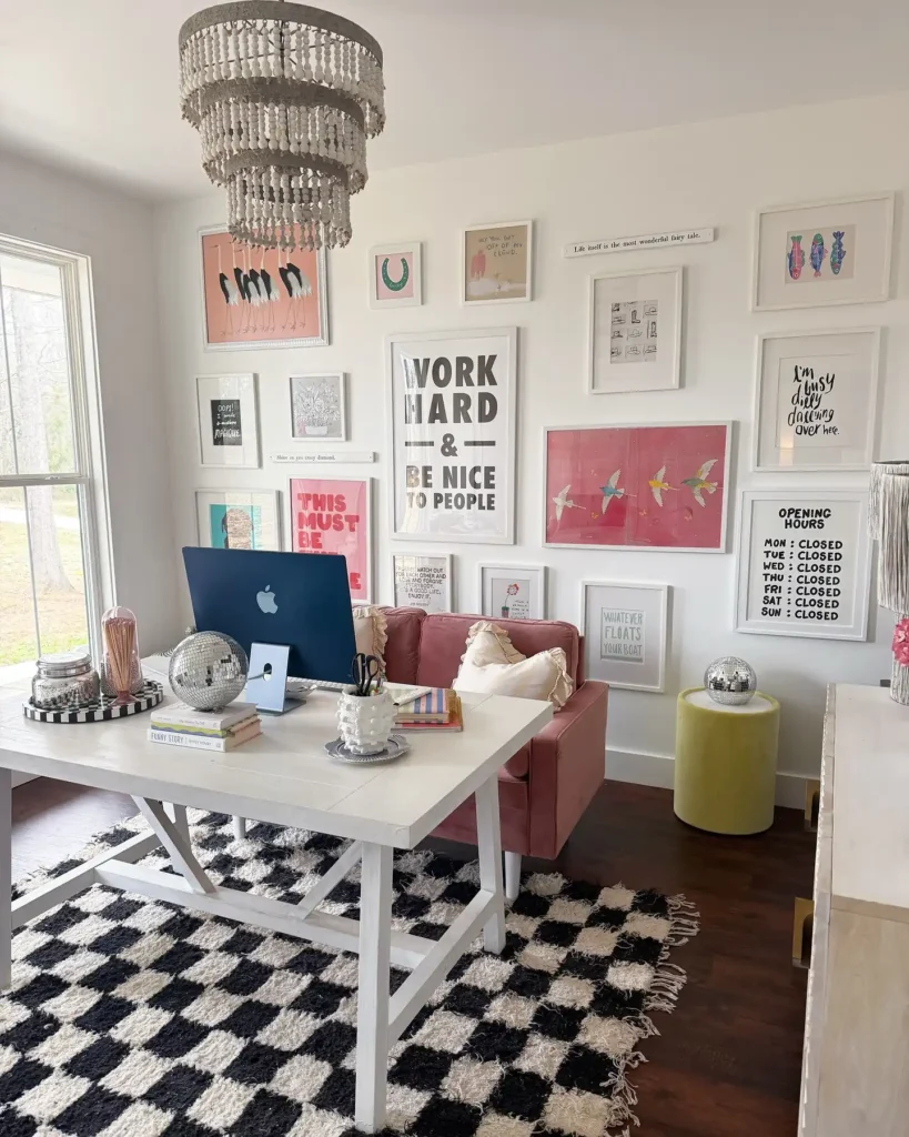 A home office with a white desk, computer, pink chair, and a gallery wall of decorative art. A black-and-white checkered rug adds style, while a large window provides natural light and a chandelier hangs above.
