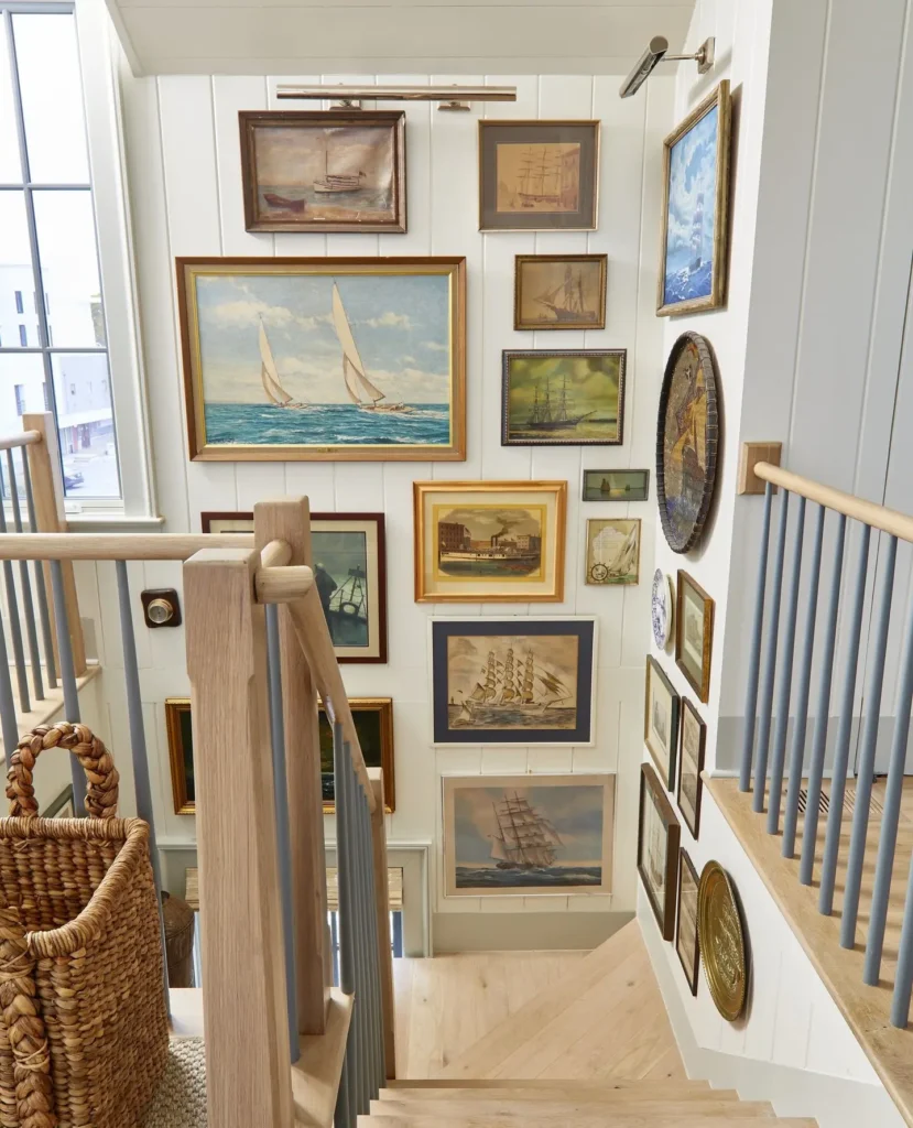 A gallery wall of framed nautical-themed paintings and prints is arranged on a white wall along a wooden staircase with gray railings and a woven basket at the bottom.
