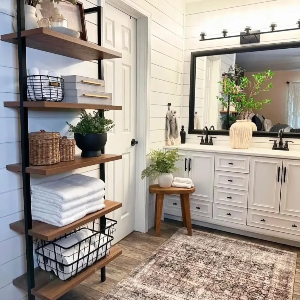 Industrial farmhouse bathroom with white shaker vanity, black hardware, open wood shelving with baskets and towels, shiplap walls, greenery accents, and a warm rustic modern bathroom design.