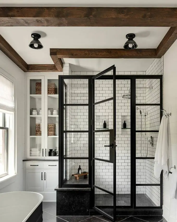 Industrial farmhouse bathroom featuring a black steel-framed glass shower, white subway tile walls, exposed wood ceiling beams, freestanding tub, and a modern rustic bathroom design.