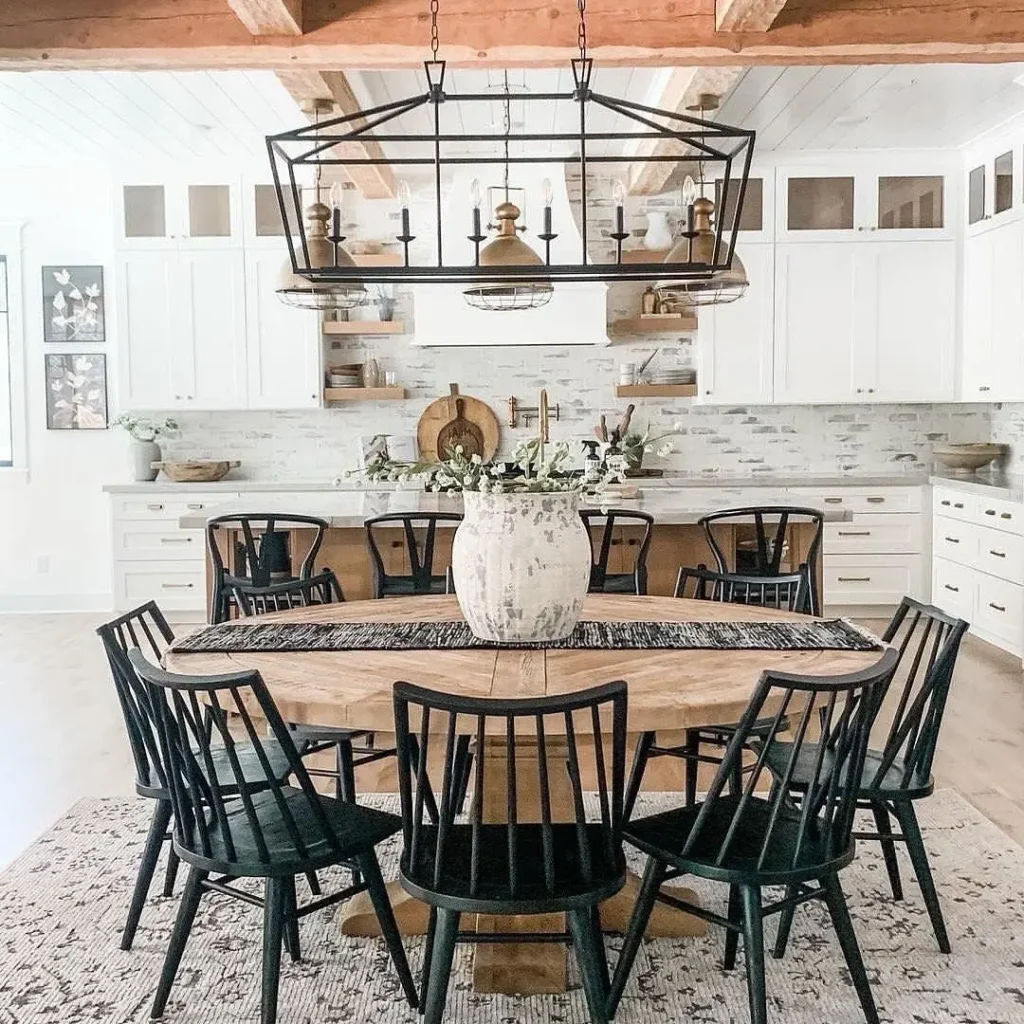 A modern industrial farmhouse dining kitchen with a wooden table, eight black chairs, a large white vase centerpiece, and a rectangular chandelier overhead. White cabinets and open shelves complete the stylish look.