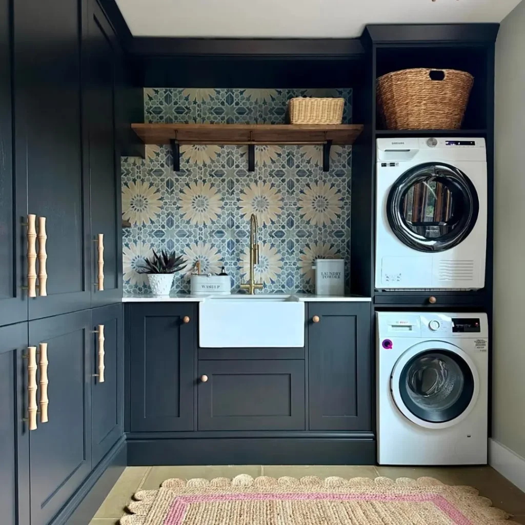 Modern laundry room with dark cabinetry, patterned tile backsplash, farmhouse sink, stacked washer and dryer, open wood shelving, and stylish built-in storage design.