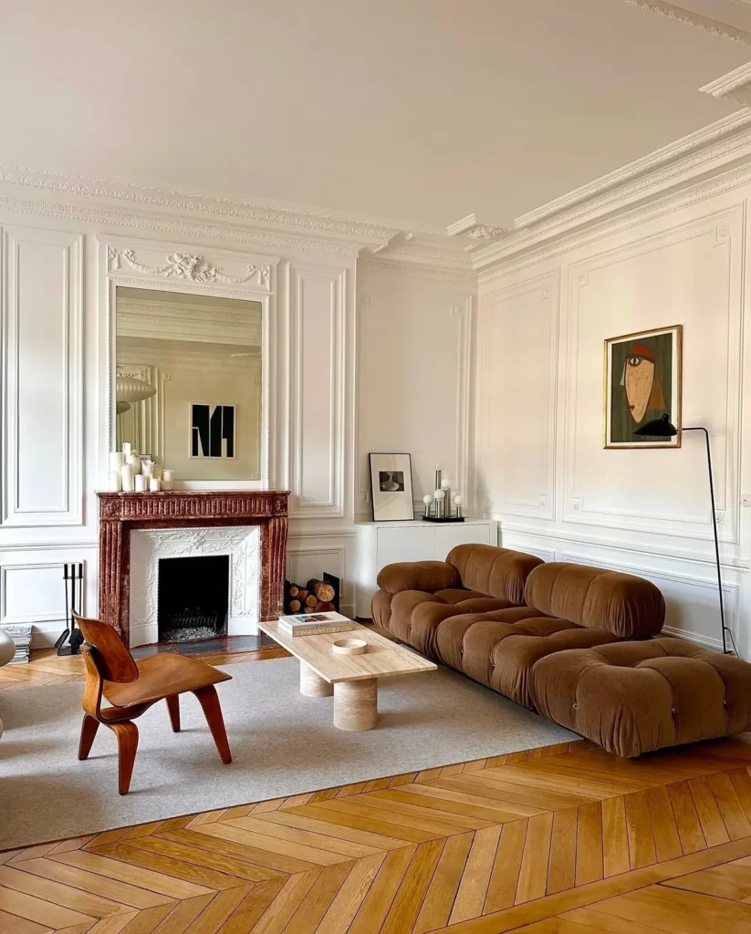 A living room with a brown tufted sofa, wooden chair, marble fireplace, and minimalist art embraces mid-century modern style, with a wooden coffee table on a beige rug atop parquet floors and ornate white walls.