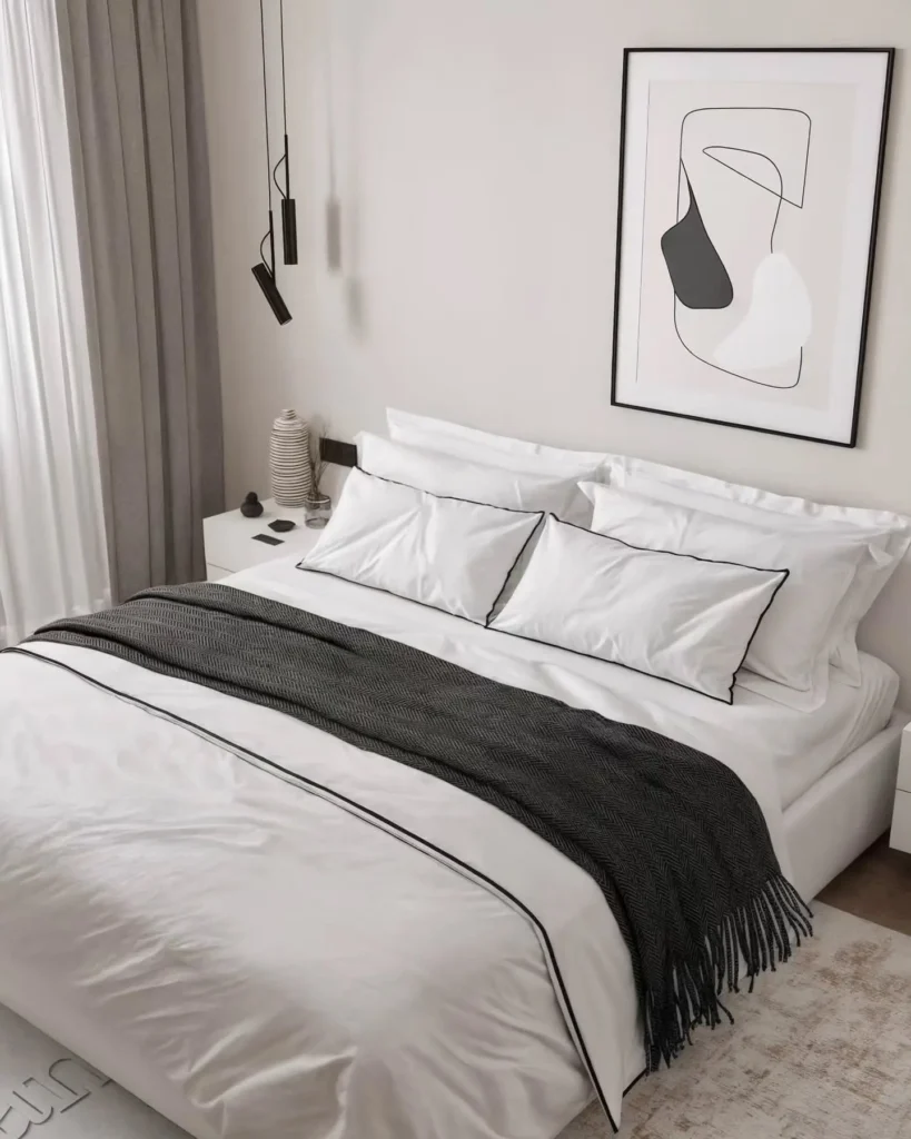 A neatly made bed with white bedding and black trim, a dark throw blanket, two nightstands, modern pendant lights, and abstract wall art create a serene minimalist bedroom.
