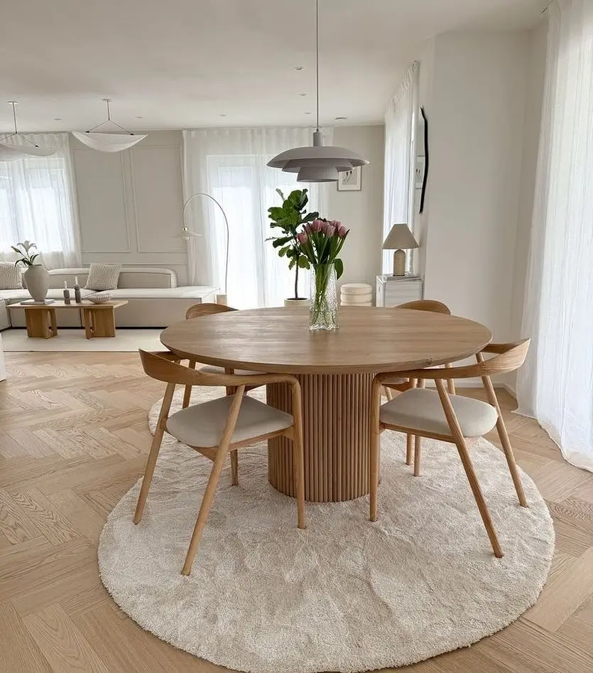 A round wooden dining table with four chairs sits on a circular rug in a bright, minimalist living space with light wood floors and simple decor. A vase with flowers adds a touch of elegance to the scene.