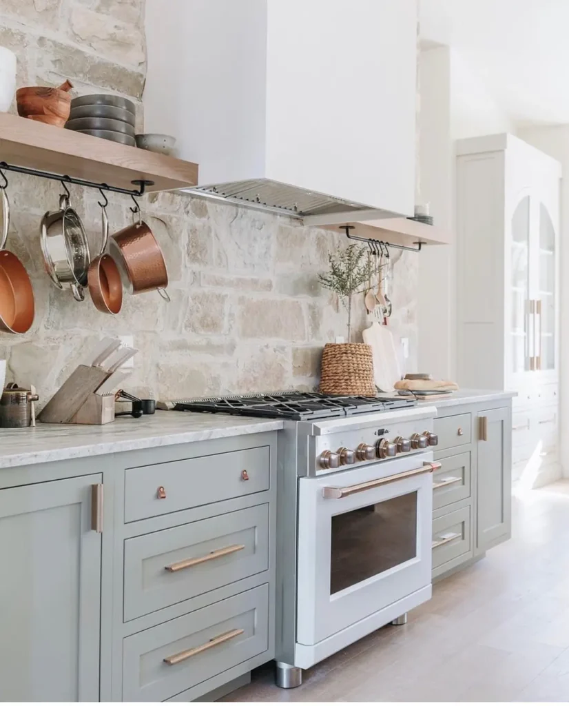 Modern cottage kitchen with light gray cabinets, a white oven, stone backsplash, wooden shelves, and copper pots hanging above the stove. Natural light brightens the space.
