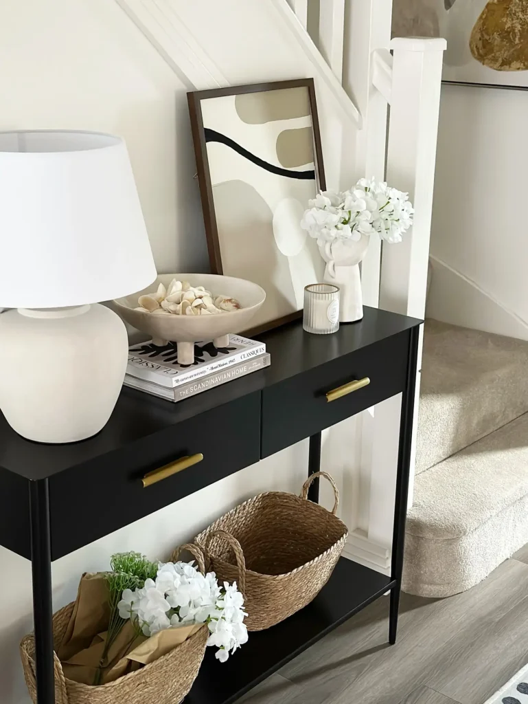 A modern black console table with two drawers holds a white lamp, books, decorative tray, framed abstract art, and vase with white flowers; baskets sit on the lower shelf by a staircase.