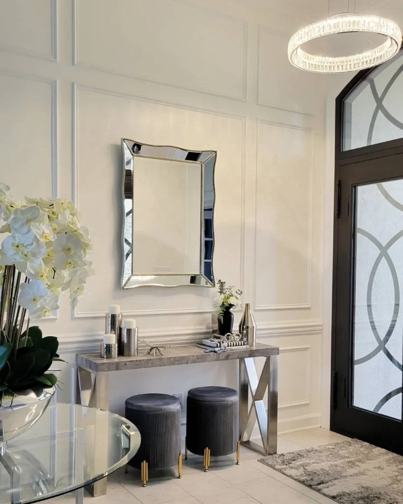 Modern entryway featuring a mirrored console table, two modern gray stools, a wall mirror, decorative vases, white orchids, glass door, and a round chandelier.