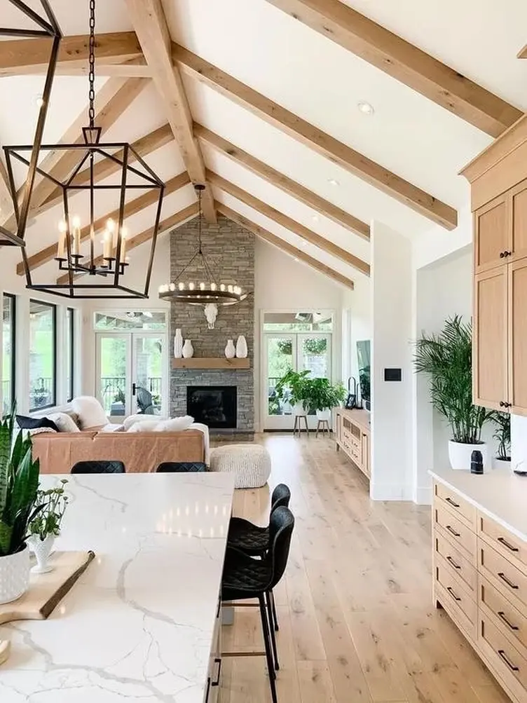 Open-concept living space with vaulted rustic wood-beam ceiling, stone fireplace, modern chandelier, kitchen island, and natural light from large windows. Green plants and neutral decor throughout.