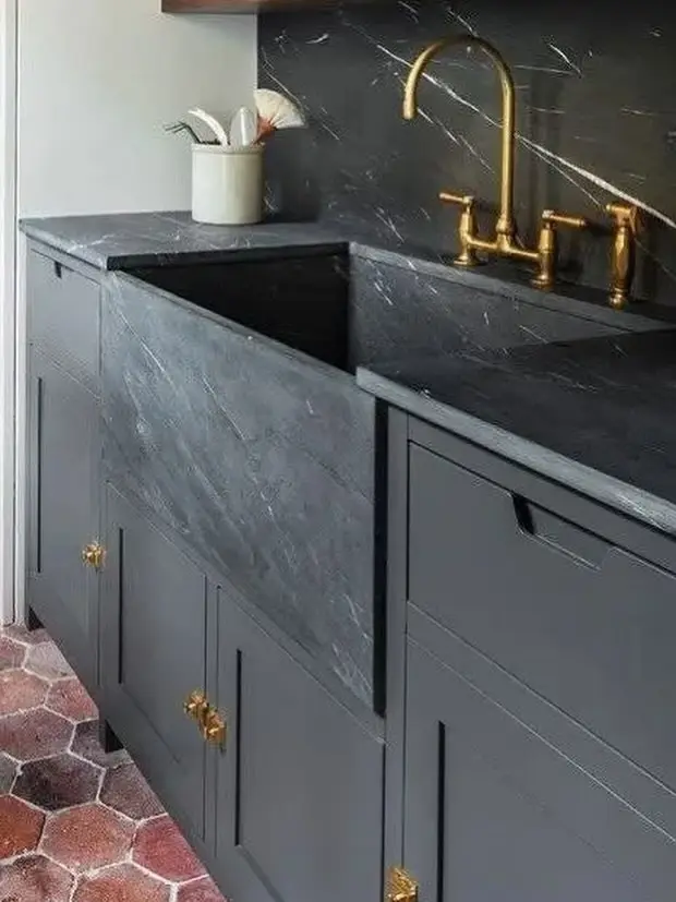A moody modern laundry featuring a dark gray farmhouse sink, matching cabinets, brass faucet, and hexagonal terracotta floor tiles.