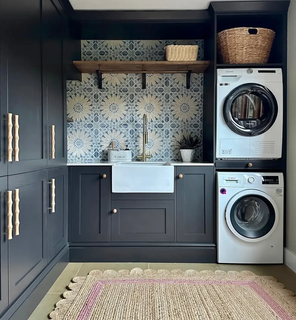 A modern laundry room with moody dark cabinets, a farmhouse sink, floral tile backsplash, stacked washer and dryer, natural wood accents, and a stylish floor rug.