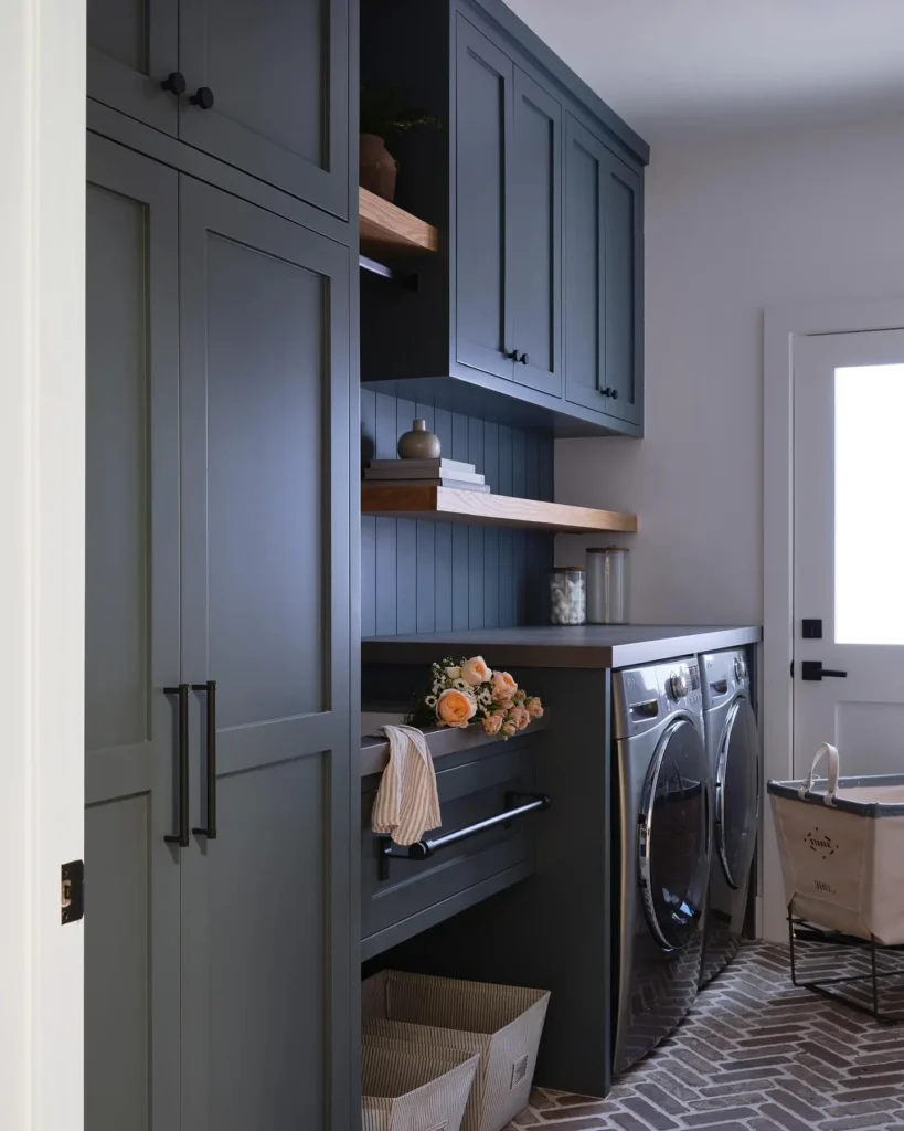 Modern laundry room with moody dark cabinetry, stacked washer and dryer, open shelving with decor, a laundry basket, and natural light from a door.