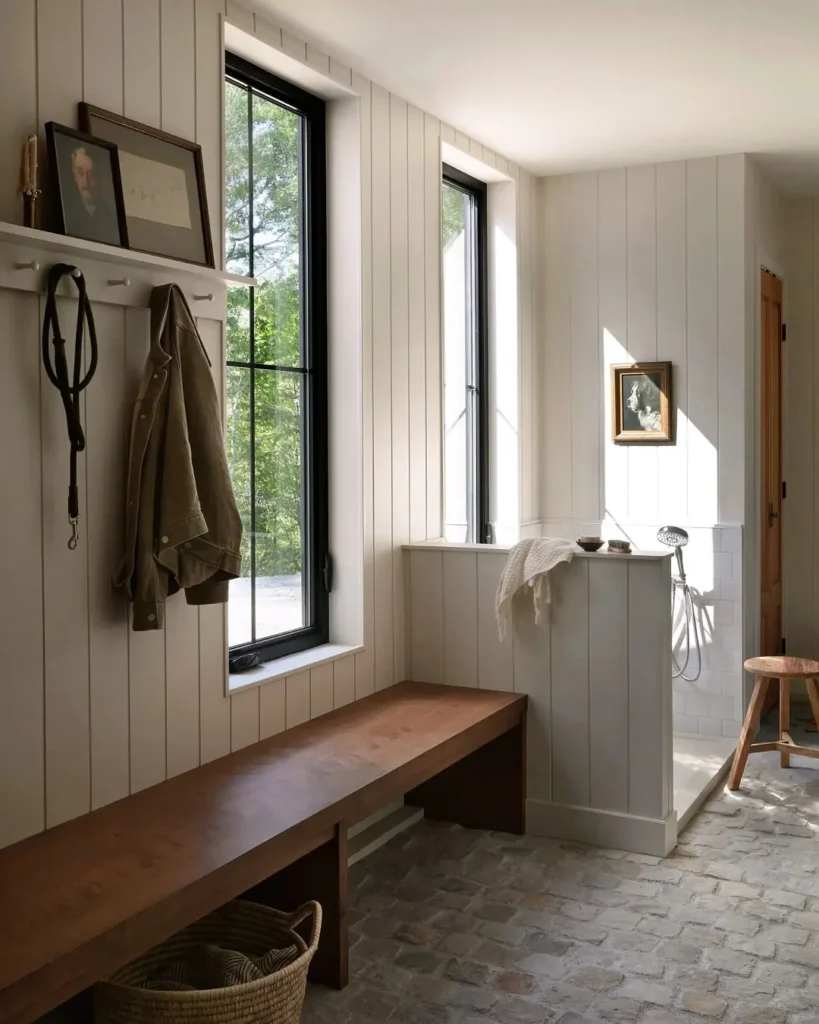 Light-filled mudroom with vertical shiplap walls, built-in wood bench, black-framed windows, wall hooks, and a walk-in pet wash area, designed for a functional and pet-friendly home entryway.