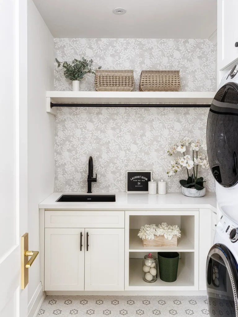 Bright laundry room with muted floral wallpaper, white cabinetry, black sink faucet, open shelving, and stacked washer and dryer.