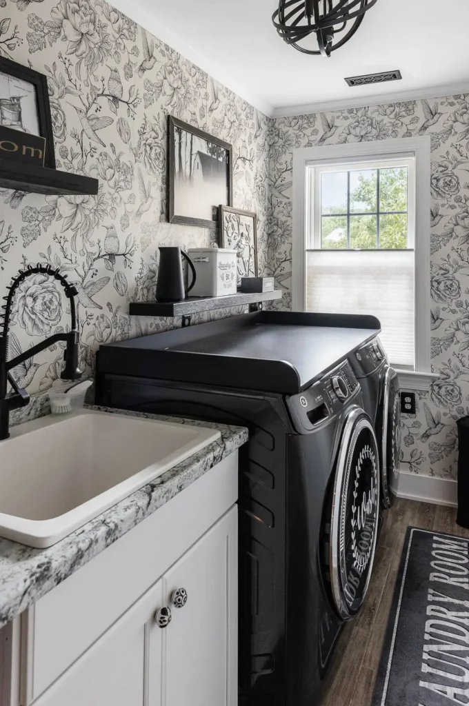 Farmhouse-style laundry room with black washer and dryer, over-the-washer folding station, floral wallpaper, granite countertop with utility sink, floating shelves, and a cozy, decorative small laundry room design.